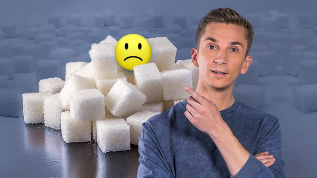 Eric Mayer im Vordergrund, dahinter jede Menge Würfelzucker. Eric zeigt mit der linken Hand auf einen kleinen Berg gestapelt aus Würfelzucker, auf dem ein negativer Smiley sitzt.