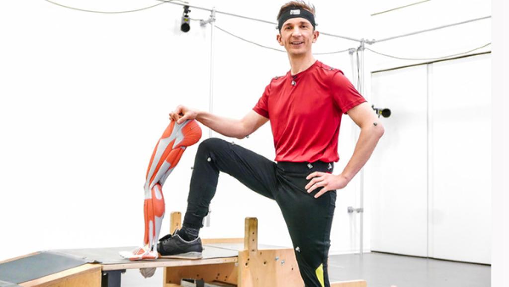 Moderator Eric Mayer steht in Sportkleidung in einem Gymnastikraum. Er hat sein rechtes Bein auf einem Turngerät aufgestützt und hält neben sein Bein das Modell eines künstlichen Beins.