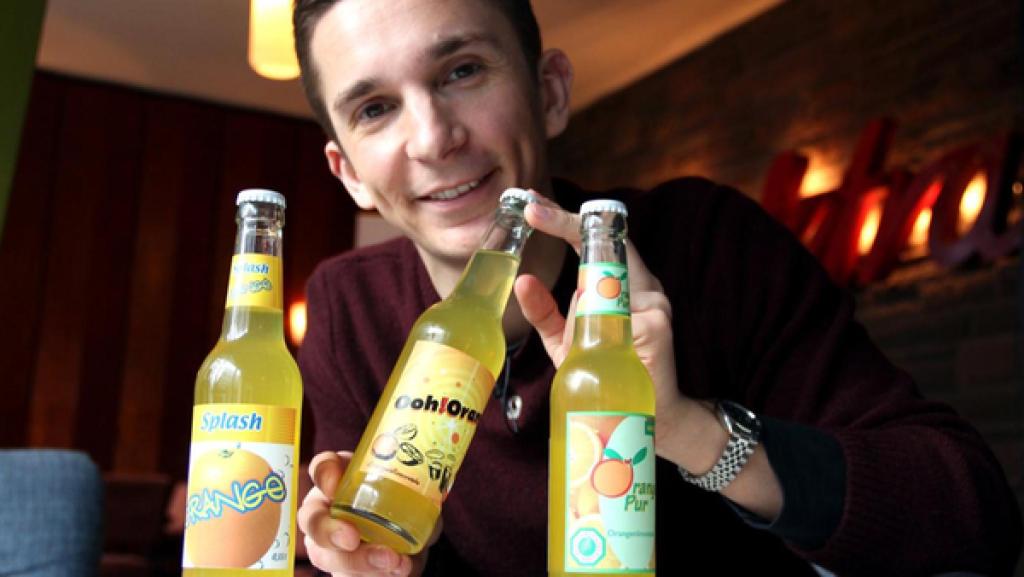 Moderator Eric Mayer hält drei Limonaden-Flaschen mit drei verschiedenen Designs in die Kamera.