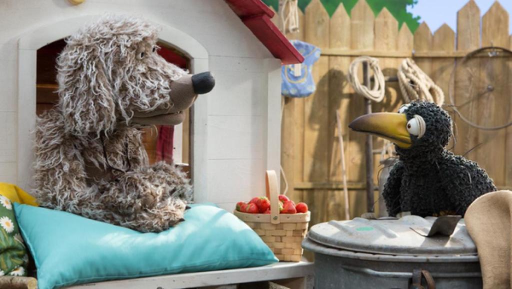 Hofhund Buffo, der in seiner Hütte hockt, und Rabe Rudi schauen auf einen Korb voller Erdbeeren, der vor ihnen steht.