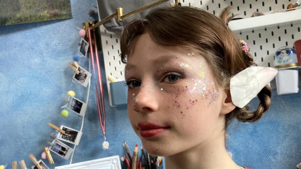 Rechte: rbb/Stefanie Köhne Ein Porträt von Julia. Sie ist mit Glitter auf Wangen und Nase als Elfe geschminkt. Ihre Haare sind geflochten und hochgesteckt. Im Hintergrund eine blaue Wand mit einer Fotogirlande und einem weißen Schreibtischboard.