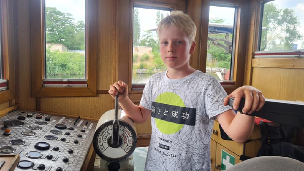Friedrich steht in einem Steuerhaus eines Schlepperbootes am Steuer. Er hat blonde kurze Haare und trägt ein weißes T-Shirt.