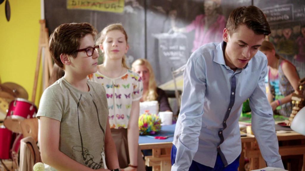 Ein Junge stützt sich auf einen Tisch, ein anderer sieht ihn von der Seite an. Dahinter sind weitere Jungen und Mädchen an Tischen.