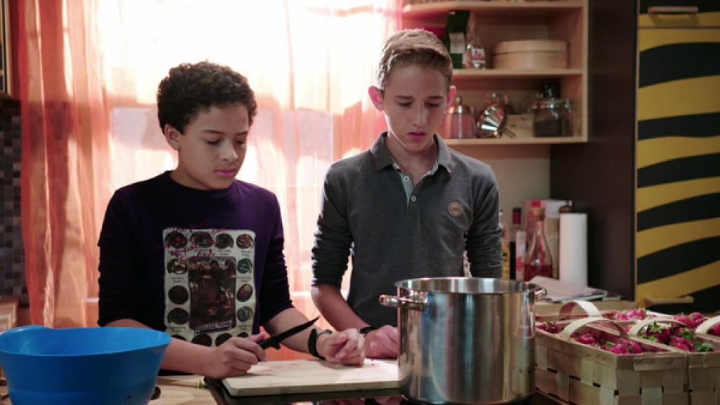 Zwei Jungen stehen an einem Küchentisch mit einem hohen Topf und Körben mit Erdbeeren daneben.