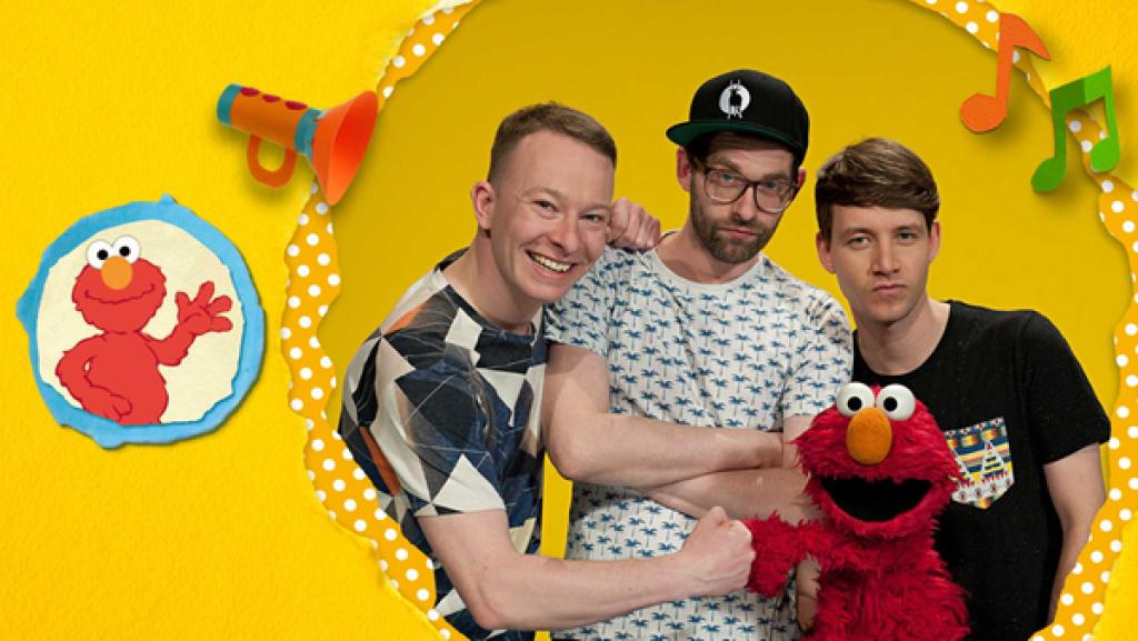 Die Band "Deine Freunde" und Elmo stehen vor einem gelben Hintergrund. Florian und Elmo begrüßen sich mit einem Faustgruß.