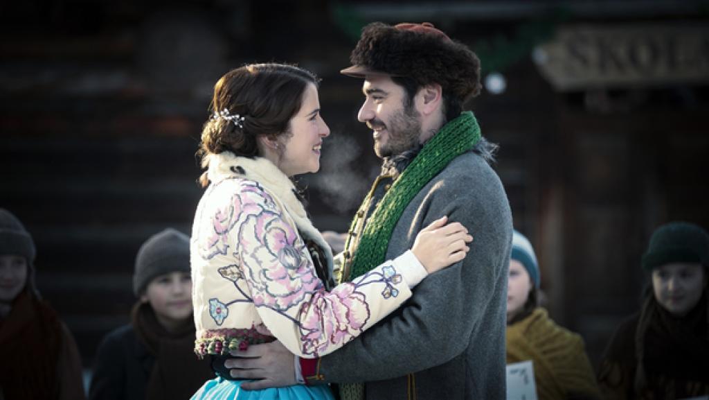 Prinzessin Amelia und Vašek stehen sich mittig gegenüber und haben ihre Arme umeinander gelegt. Sie schauen sich glücklich in die Augen. Im Hintergrund ein Holzhaus und Kinder, die zu ihnen schauen.