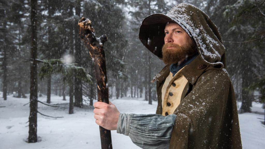 Krakonos steht mit Umhang und hochgezogener Kapuze in einem verschneitem Wald. Er stützt seinen Arm auf einen großen Wanderstock aus Holz. Sein Blick ist ernst.