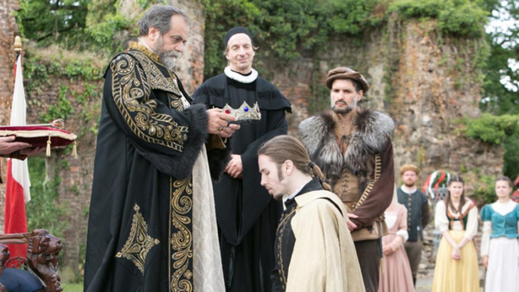 Ein Mann im Gewand setzt einem jüngeren Mann, der vor ihm kniet, eine Krone auf. Im Hintergrund beobachten zwei weitere Männer die Szene