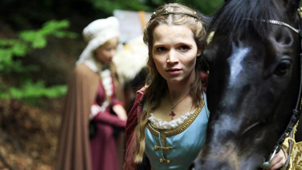 Prinzessin Elisabeth steht in einem dämmrigen Wald. Dicht neben ihr ist ein Pferdekopf von einem schwarzen Rappen. Elisabeth trägt ein blaues Gewand und hat lange blonde Haare. Im Hintergrund, ist eine weiter Frau in einem braunen Kleid mit weißer Haube zu sehen.