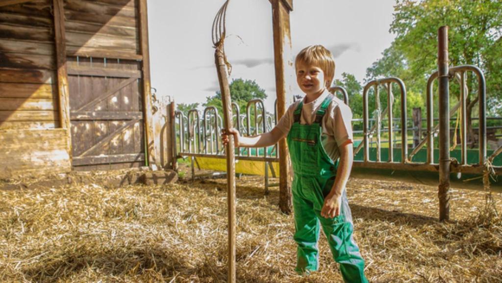 Justus steht im Heu mit einer Mistgabel in der Hand und lacht.