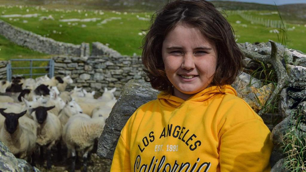 Lena lächelt freundlich in die Kamera. Sie hat halblange, dunkle Haare und trägt einen knallgelben Hoodie. Sie steht in einem Schafsgatter, das von Steinen begrenzt ist. Neben ihr drängen sich etliche Schafe zusammen. Im Hintergrund kann man schottische Weidelandschaft erkennen.
