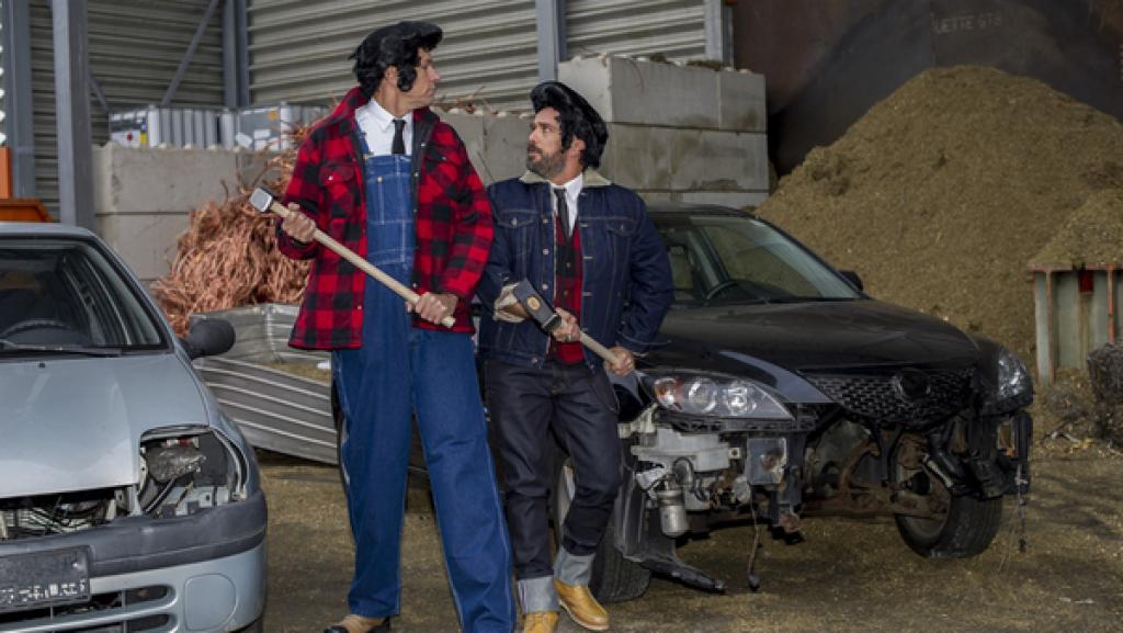 Zwei Männer hämmern abwechselnd im Takt auf ein Autowrack ein.
