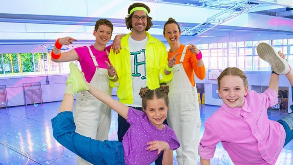 Die TanzAlarm Kids stehen in sportlichen Posen vor Kindermusiker herrH und seinen Tänzerinnen mitten in der Sporthalle. TanzTapir steht rechts an der Seite mit einem Kletterseil in der Hand.