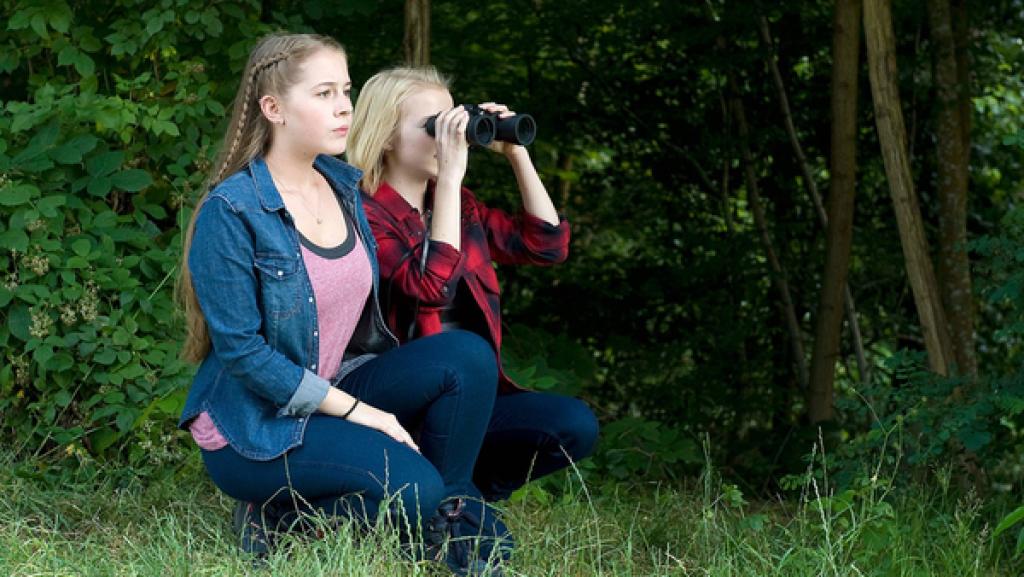 zwei Mädchen ducken sich am Wald und beobachten durch ein Fernglas