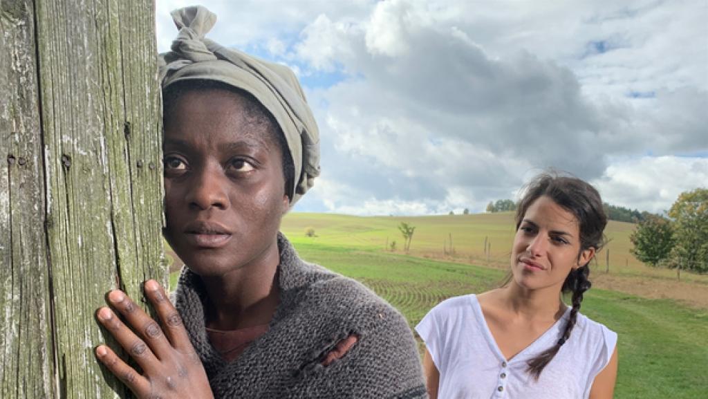 Rechte: KiKA/Sabine Finger Clarissa steht hinter Harriet Tubman (Nancy Mensah-Offe) auf einem Feld. Tubman lehnt mit Körper und Gesicht an einen Holzphal. Sie schaut lauschend geradeaus. Clarissa beobachtet sie.
