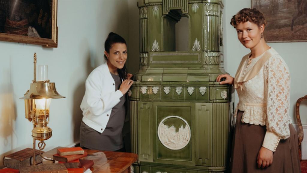 Rechte: KiKA/ Sabine Finger Clarissa und Bertha stehen links und rechts neben einem großen Kachelofen in einem Zimmer und lächeln. Links ist ein kleiner Tisch mit Büchern und einer Lampe. An den Wänden sind zwei Gemälde.