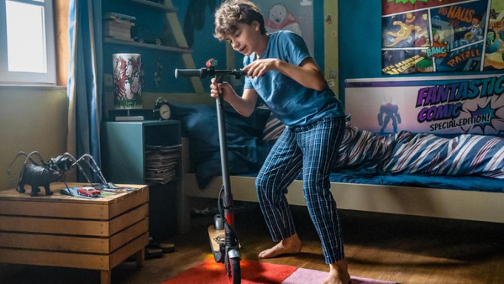 Jans Zimmer in der ersten Morgensonne: Plakate an den Wänden, im Hintergrund das Bett, links ein Holzschränkchen, auf dem Boden ein bunter Teppich. Darauf steht – im Schlafanzug – Jan (mitte) und umfasst mit beiden Händen, voller Begeisterung, das Lenkrad eines edlen, voll funktionsfähigen und sogar mit einer Lampe ausgestatteten Scooters.