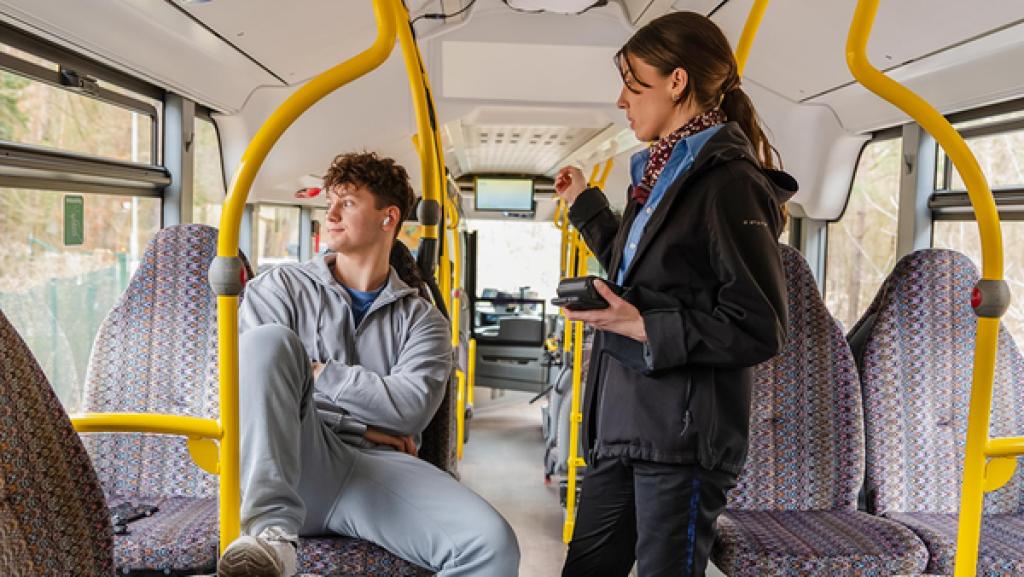 Young Crime - Staffel 2 - Unverschämt und ohne Ticket | Rechte: ZDF Im hinteren Teil eines Busses sitzt ein Jugendlicher im grauen Jogginganzug. Er hat seine Arme verschränkt und seinen rechten Fuß auf dem Sitz vor ihm abgestellt. Trotzig und genervt schaut er aus dem Fenster und ignoriert die Frau, die neben ihm steht. Sie trägt eine dunkle Jacke und eine blaue Bluse und schaut ihn tadelnd an. In der linken Hand hält sie zwei kleine Geräte zur Ticketkontrolle, den Zeigefinger der rechten Hand hat sie streng erhoben und zeigt in Richtung Ausgang des Busses.